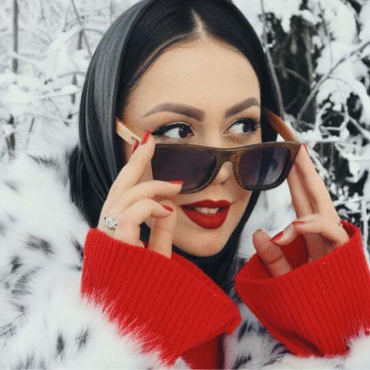 Stilvolle Frau mit rotem Lippenstift und Sonnenbrille im Winter-Outfit vor verschneiter Kulisse – eleganter Look mit Statement-Ring und Fellmantel.