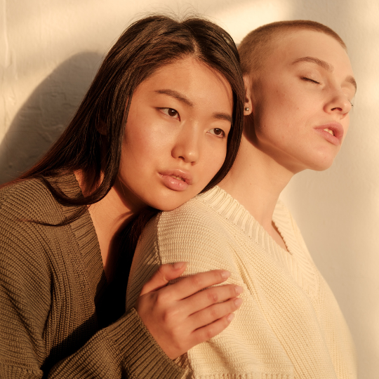 Eine asiatische junge Frau umarmt von hinten eine europäische Frau mir kurz geschorenen Haaren