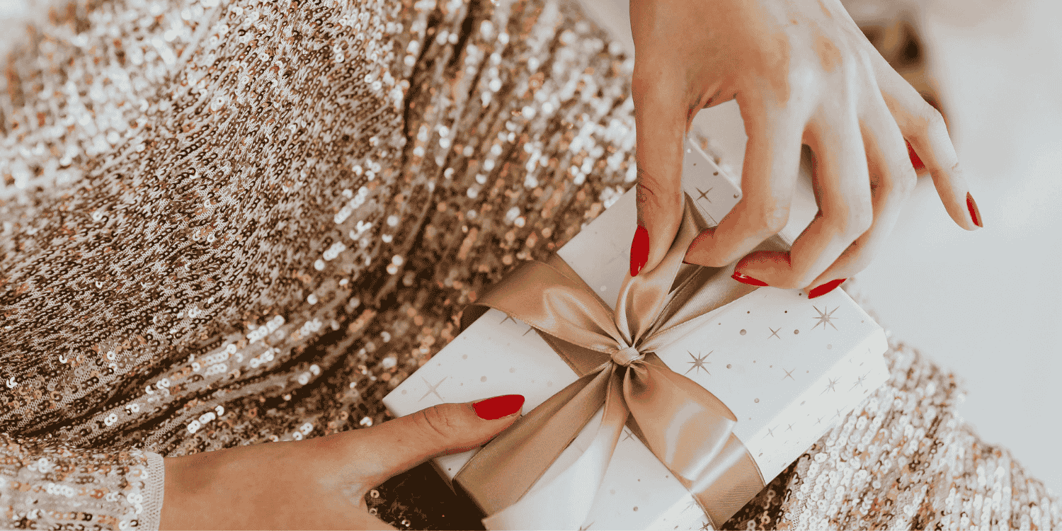 Frau im goldenen Pailettenkleif die mit auf ihrem Schoß ein Geschenk mit einer goldenen Schleife auspackt